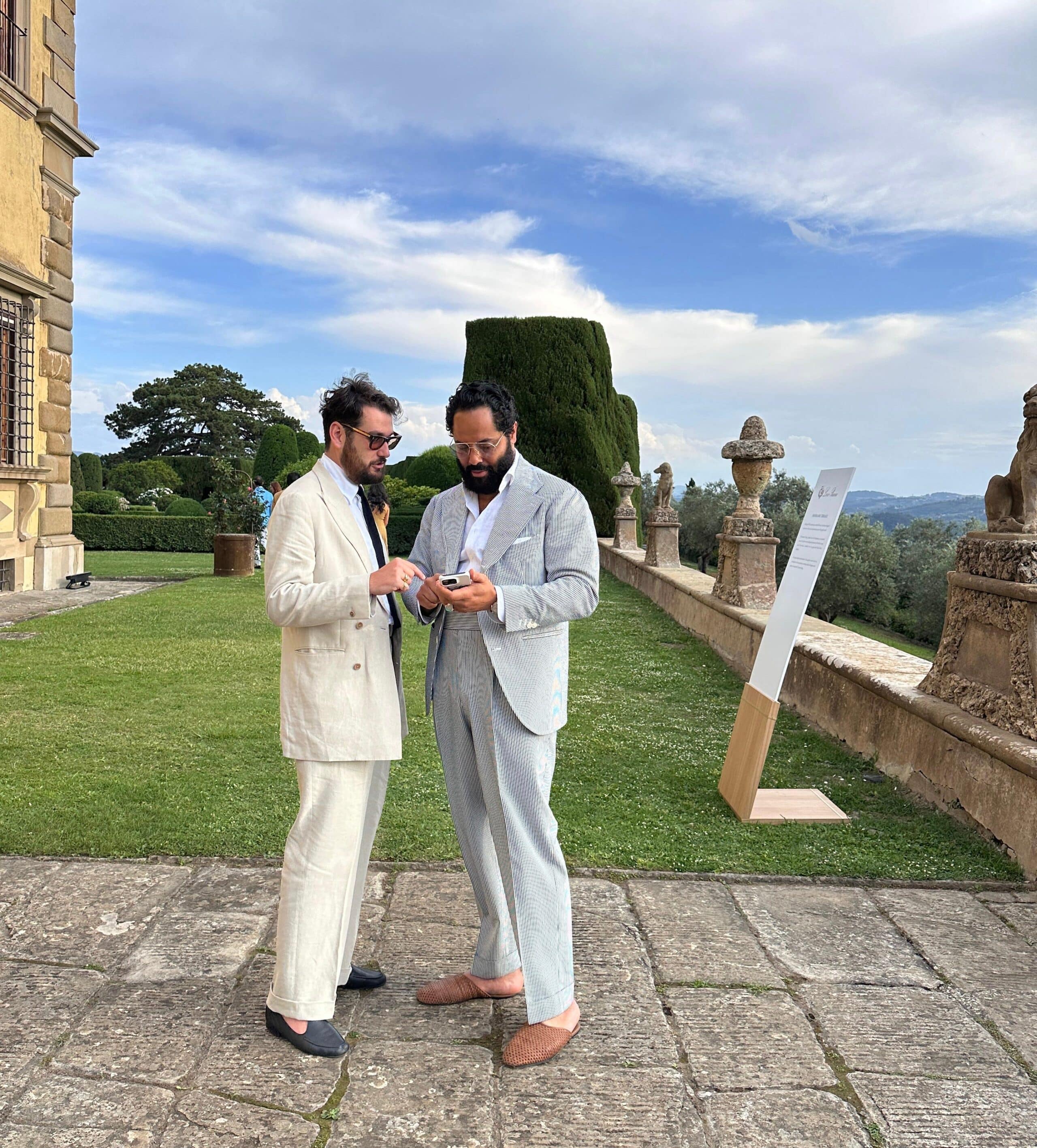 Angel Ramos and Matt Hranek stand on a stone pathway in an elegant garden, engaged in conversation while looking at a phone.