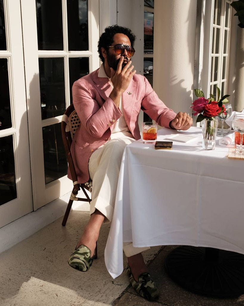 Angel Ramos in a pink blazer and white trousers enjoys a Negroni while smoking a cigar at an outdoor café.
