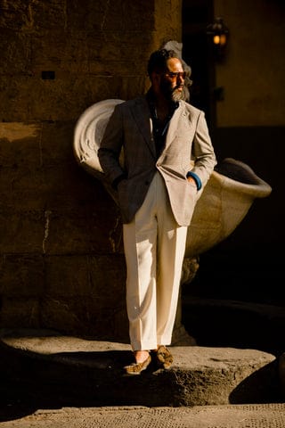 Angel Ramos in a light gray blazer and ivory trousers leaning against a historic stone fountain, bathed in golden sunlight.