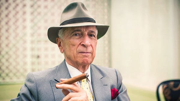 Famed journalist and author Gay Talese in a tailored gray suit and fedora, elegantly holding a cigar.