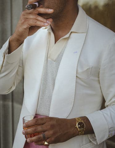 Angel Ramos in an ivory jacket and cream knit polo smokes a cigar while holding a glass of whiskey.