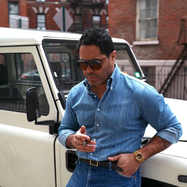 Angel Ramos wearing a denim button-down shirt and sunglasses, enjoying a cigar while leaning against a classic white Land Rover in New York City.
