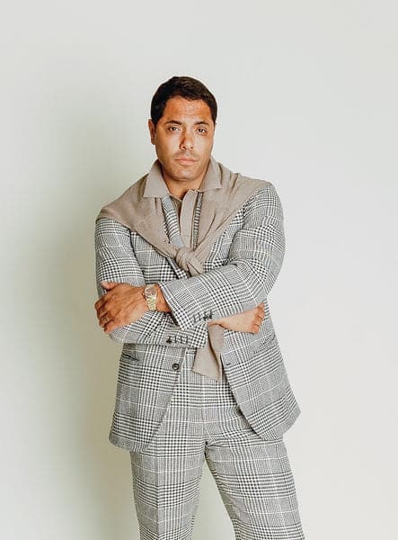 Angel Ramos wearing a summer tweed Glen plaid suit with a taupe knit polo and a matching sweater draped over his shoulders, showcasing the refined texture of raw silk.
