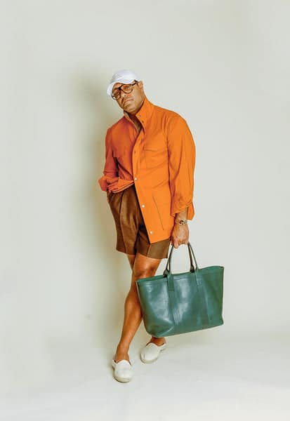 Angel Ramos wearing an orange Sahariana jacket with brown tailored shorts, white espadrilles, and a green leather tote, embracing a bold yet sophisticated summer look.