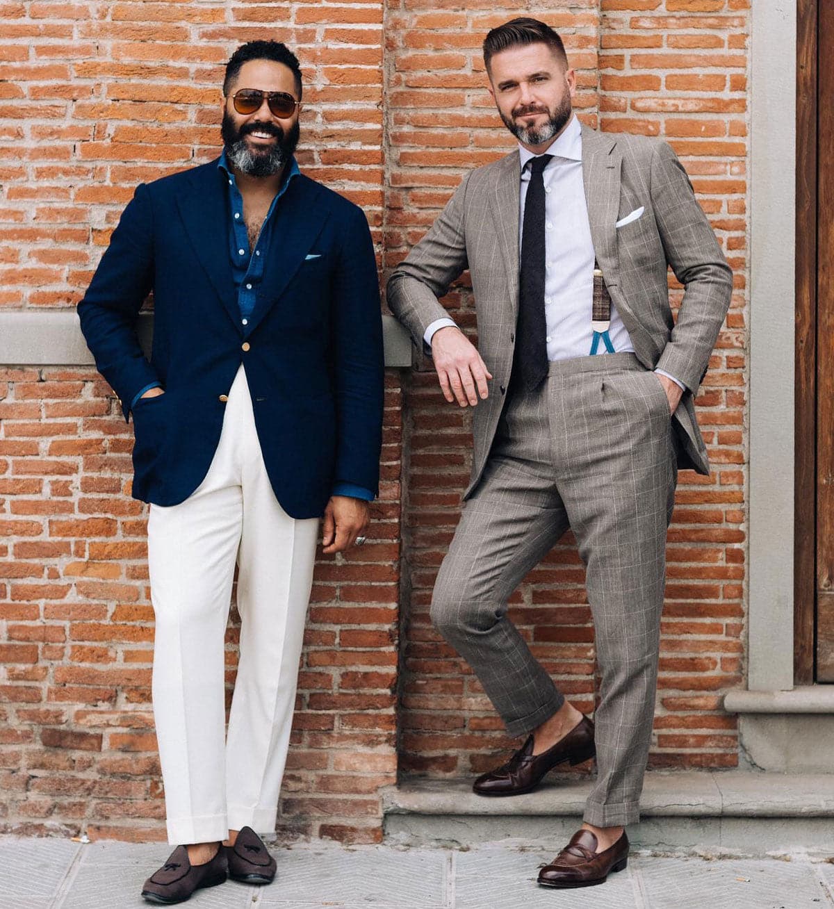 Angel Ramos and Stephen posing against a brick wall; Angel in a navy blazer with a denim shirt and white trousers, and Stephen in a light brown plaid suit with suspenders and a dark tie.